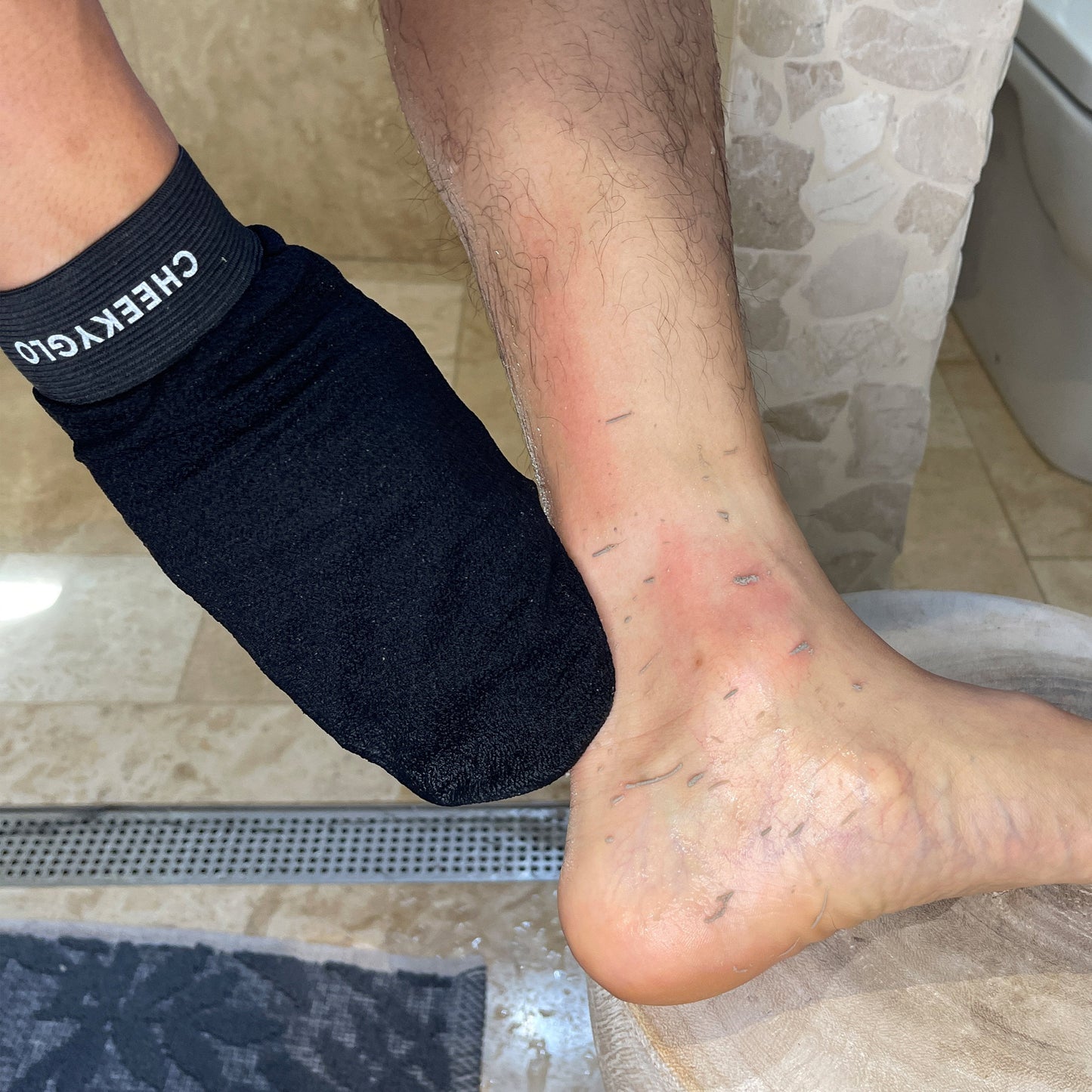 A person is exfoliating their left foot using a CheekyGlo Exfoliating Glove. The foot shows some visible signs of dry skin, redness, and small scratches. The background includes a bathroom setting with tiled walls and floor, a toilet, and a black mat. The exfoliation promises smoother skin for healthier feet.