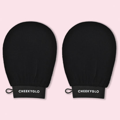 A pair of black exfoliating gloves, known as the "CheekyGlo Exfoliating Glove," features a textured surface and a loop for hanging. Displayed against a pastel pink background, these gloves are designed to help you achieve smoother skin. The brand name "CheekyGlo" is written in white on the elastic wristbands.