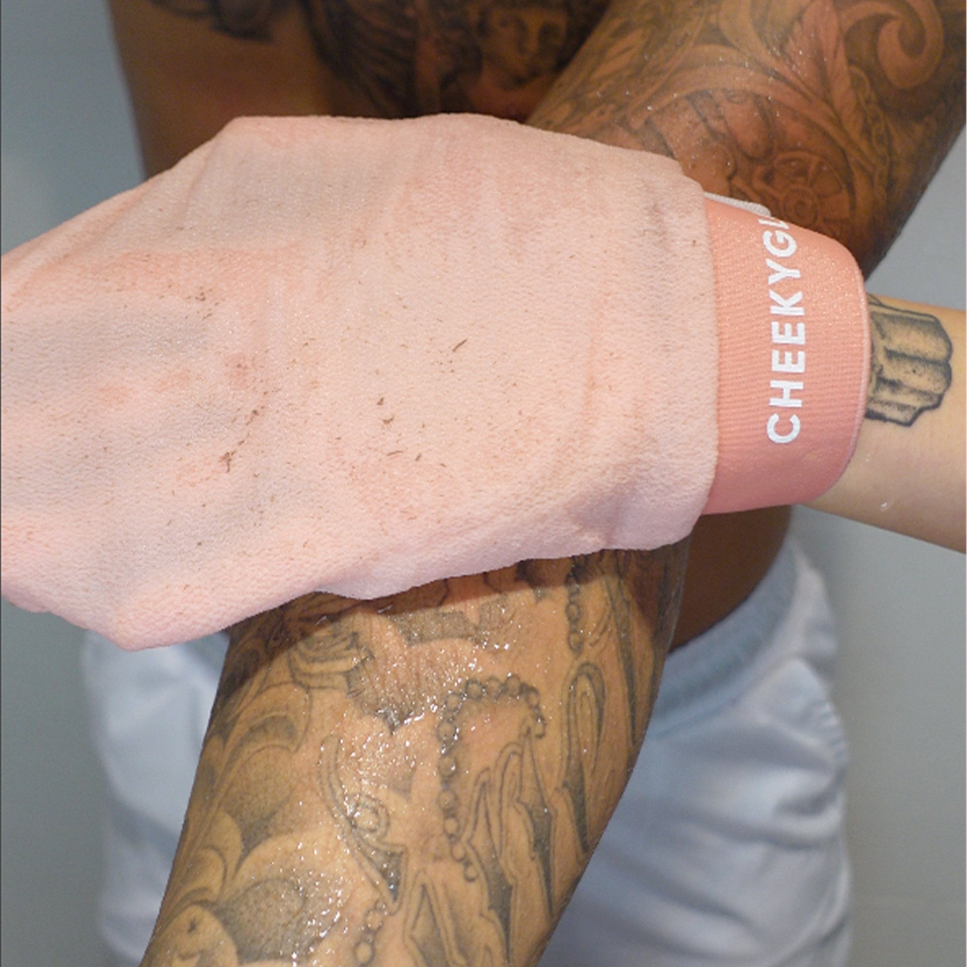 A tattooed individual, wearing white shorts, rubs their arm with a pink CheekyGlo Exfoliating Glove from the brand CheekyGlo, aiming for healthier, smoother skin.