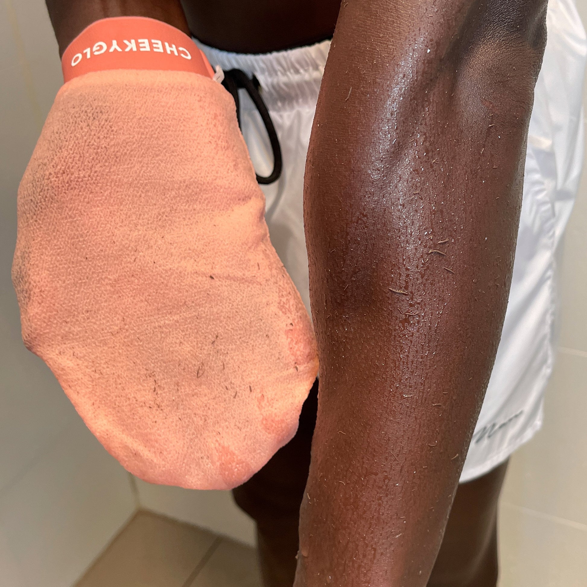 A person wearing white shorts is using a CheekyGlo Exfoliating Glove on their right arm. The light orange glove, branded "CheekyGlo" on the wristband, glides over damp skin. The bright lighting accentuates the textures on the arm and glove, promising smoother and healthier skin with each use.