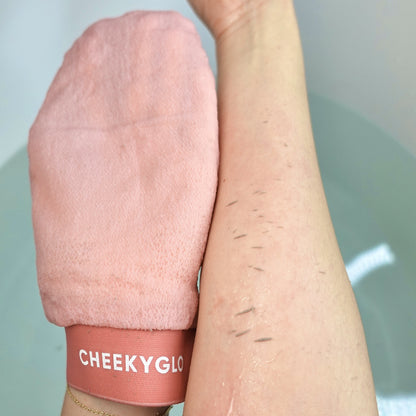 A close-up of a person's arm shows exfoliated dead skin cells being removed with a pink CheekyGlo Exfoliating Glove from CheekyGlo. The glove is gently scrubbing the forearm, revealing smoother and healthier skin underneath.