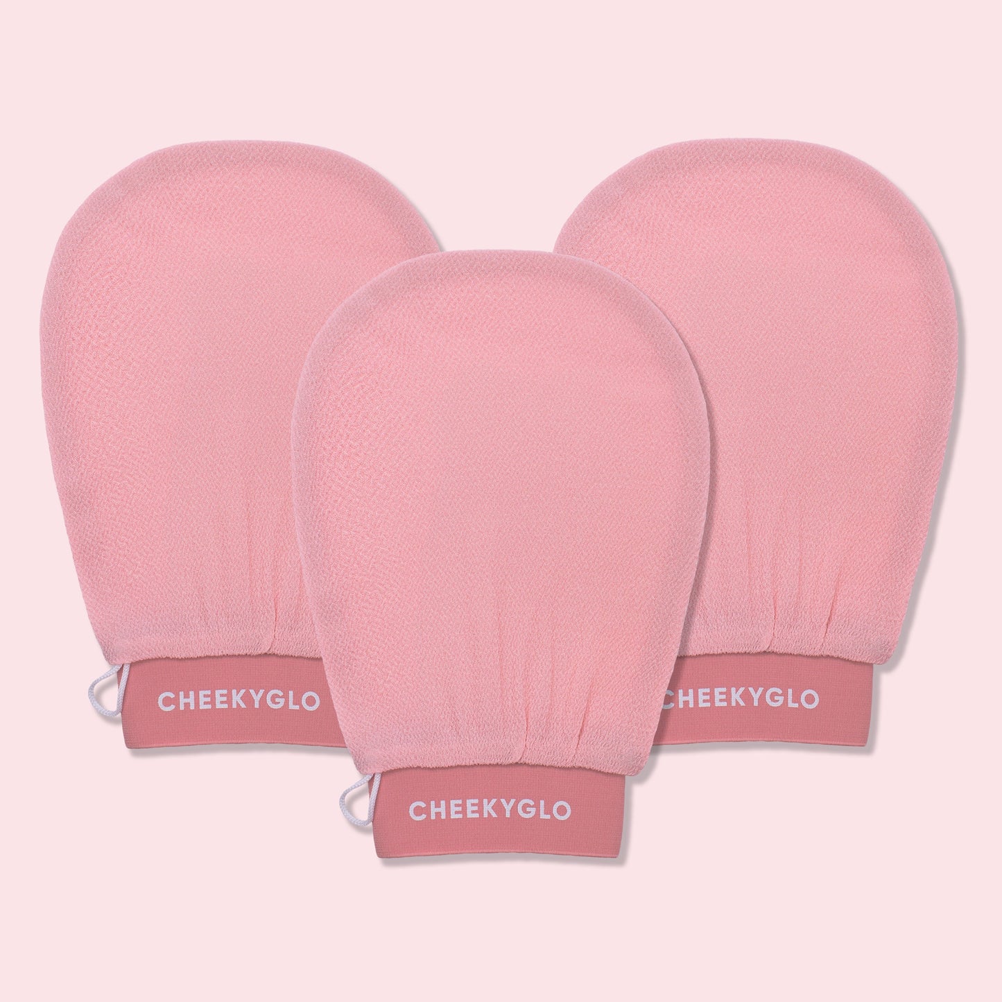 Three CheekyGlo Exfoliating Gloves are displayed against a light pink backdrop. The gloves are slightly rounded at the top with elasticated wristbands labeled "CHEEKYGLO," promising smoother skin and a healthier glow.