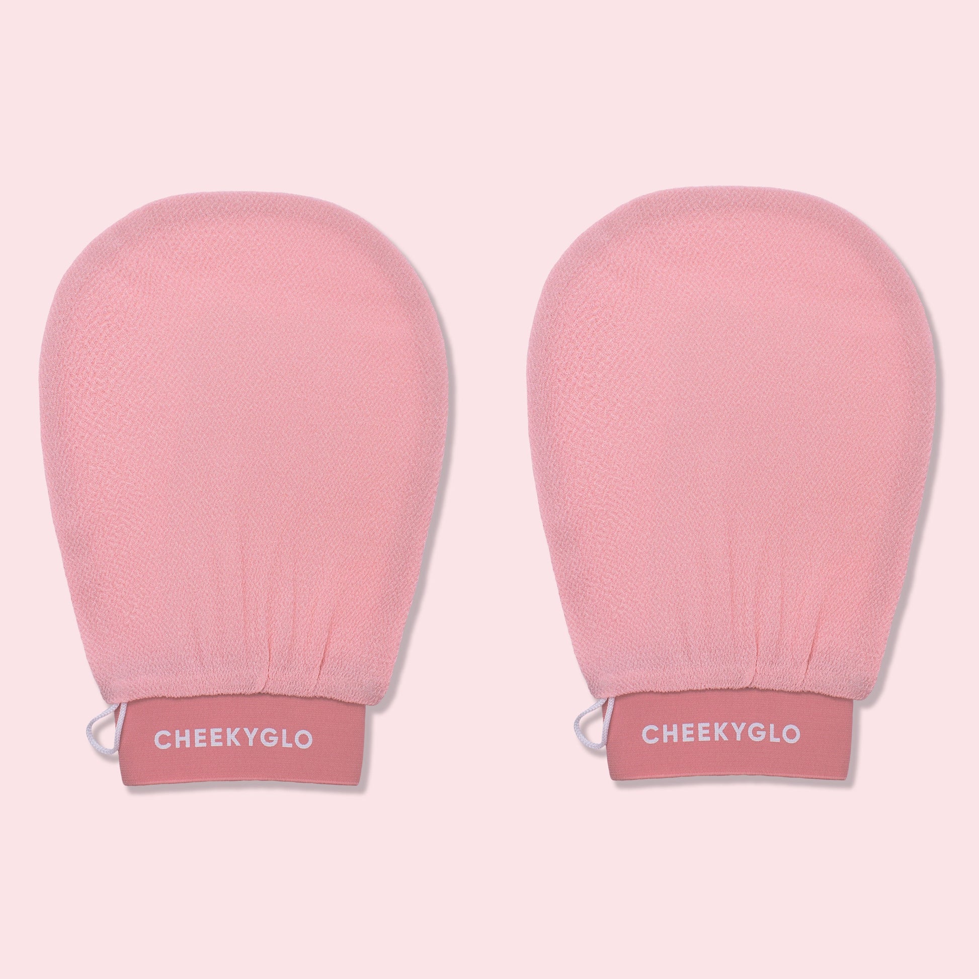 Two pink CheekyGlo Exfoliating Gloves with "CheekyGlo" printed in white on the wristbands. The gloves promise smoother, healthier skin and are set against a light pink background.